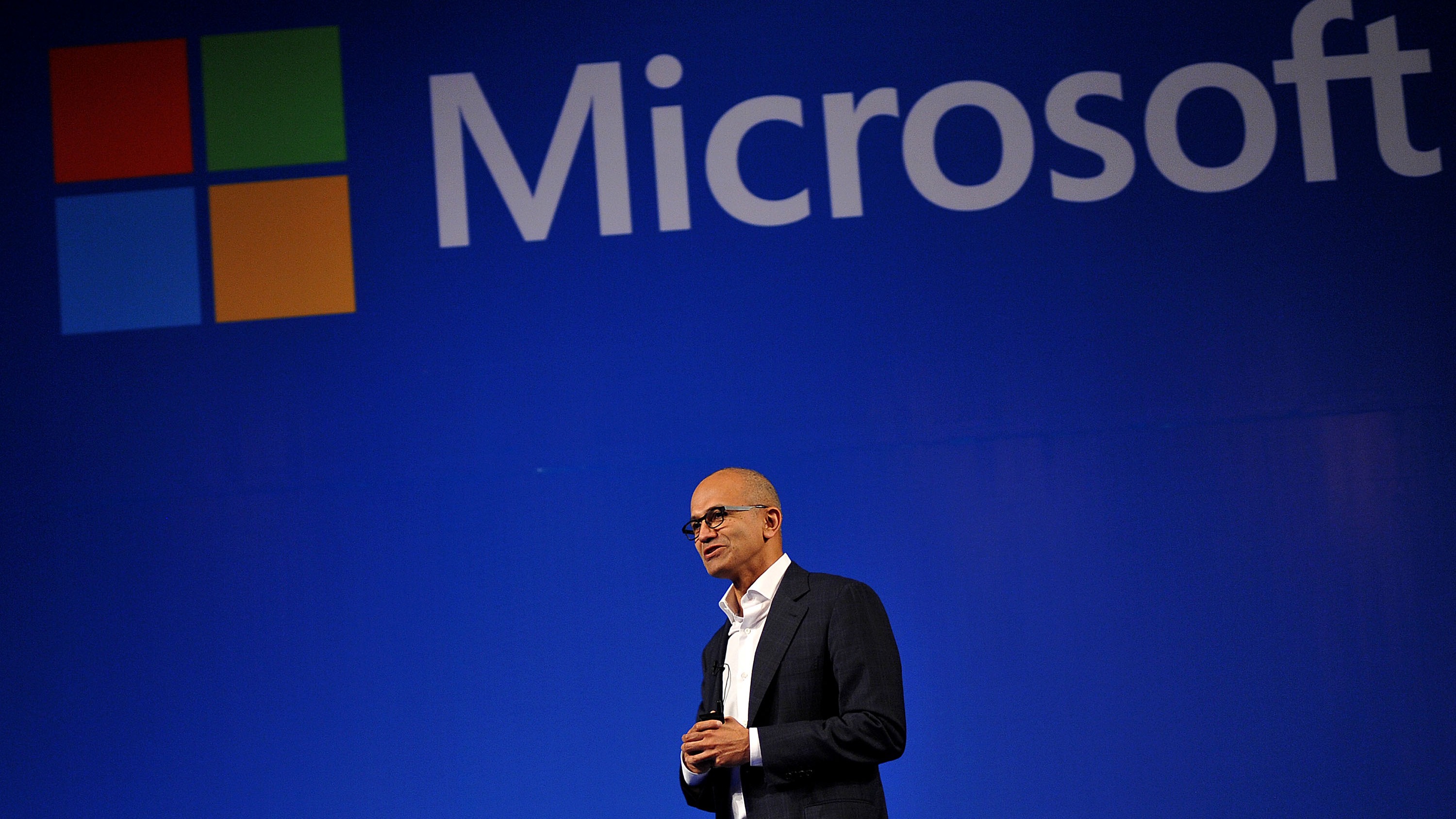 萨提亚·纳德拉在微软活动舞台上演讲，背景为蓝色背板与 Microsoft 标志