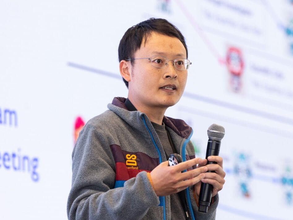 James Zou 手持麦克风讲解，身后屏幕上是智能体协作节点示意图