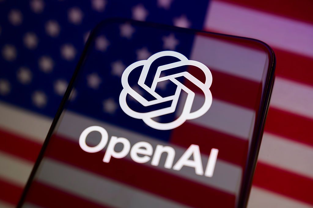 手机屏幕上的 OpenAI 标识，背景为美国国旗（TechCrunch 报道配图）