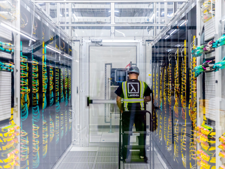 Lambda 数据中心里，一名身穿 Lambda 背心的技术人员走在 GPU 机架通道中