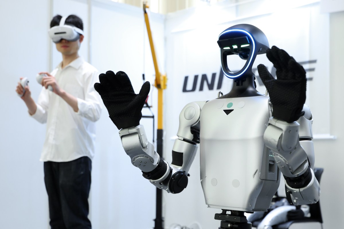 宇树科技人形机器人在演示中举起双臂，背景是佩戴 VR 头显的操作者和 UNITREE 标识（Kiyoshi Ota/Bloomberg via Getty Images，via TechCrunch）