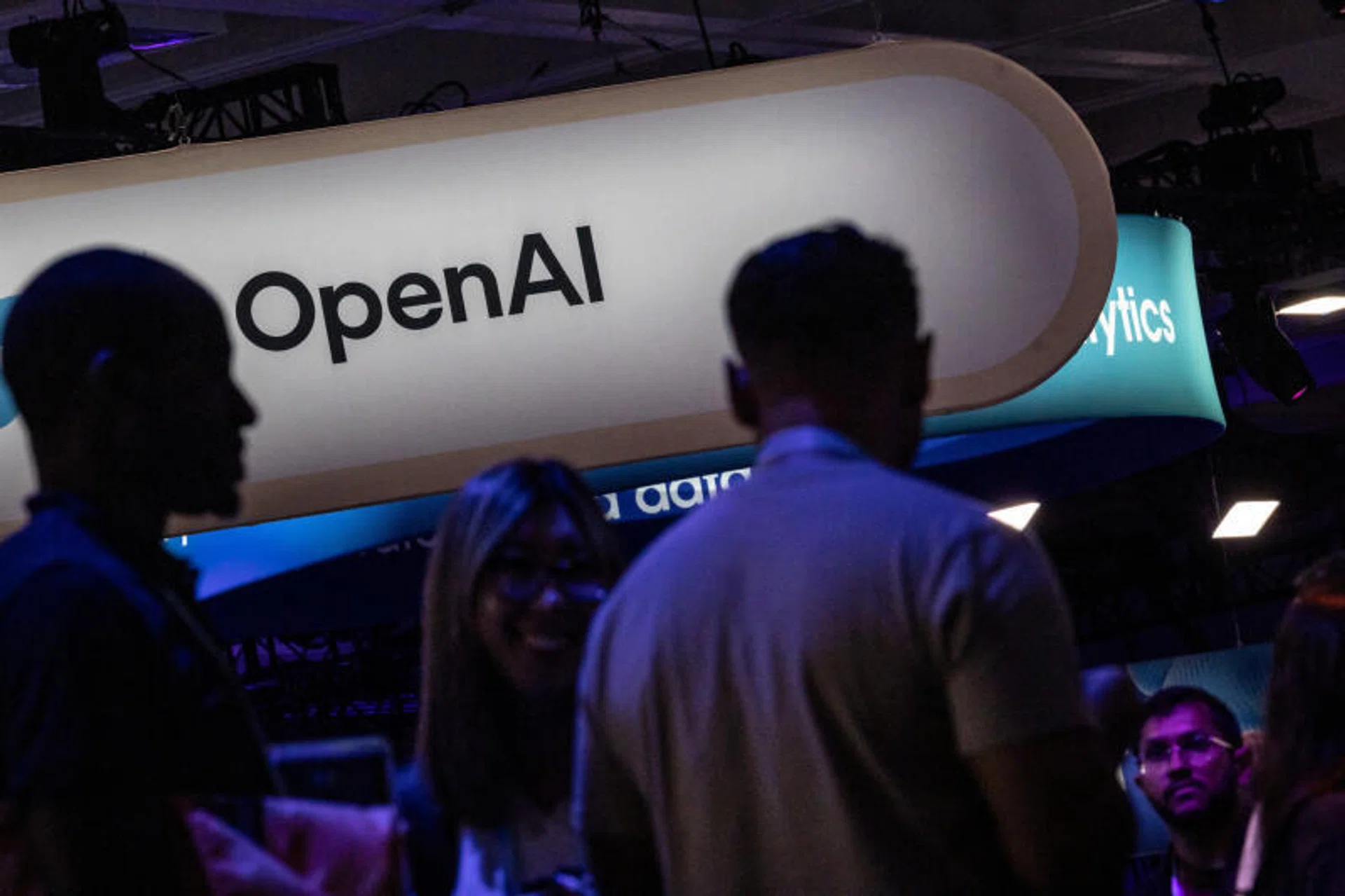 一场科技展会上，OpenAI 标志悬挂在展台上方，前景是与会者的剪影
